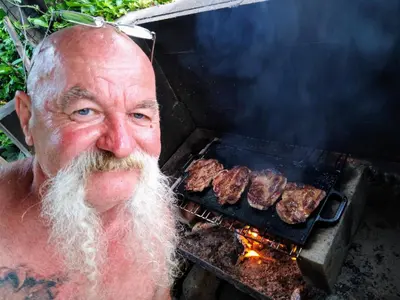 an older man with a white beard is cooking meat on a grill at Penzion Lida in Srbsko