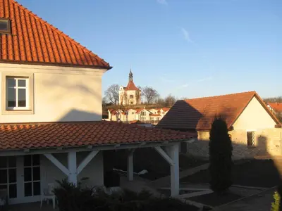 a building with an orange tiled roof with a church in the background at Penzion V polích in Malé Číčovice