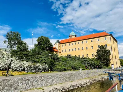 a large building with a red roof next to a river at Královské apartmá kolonáda in Poděbrady